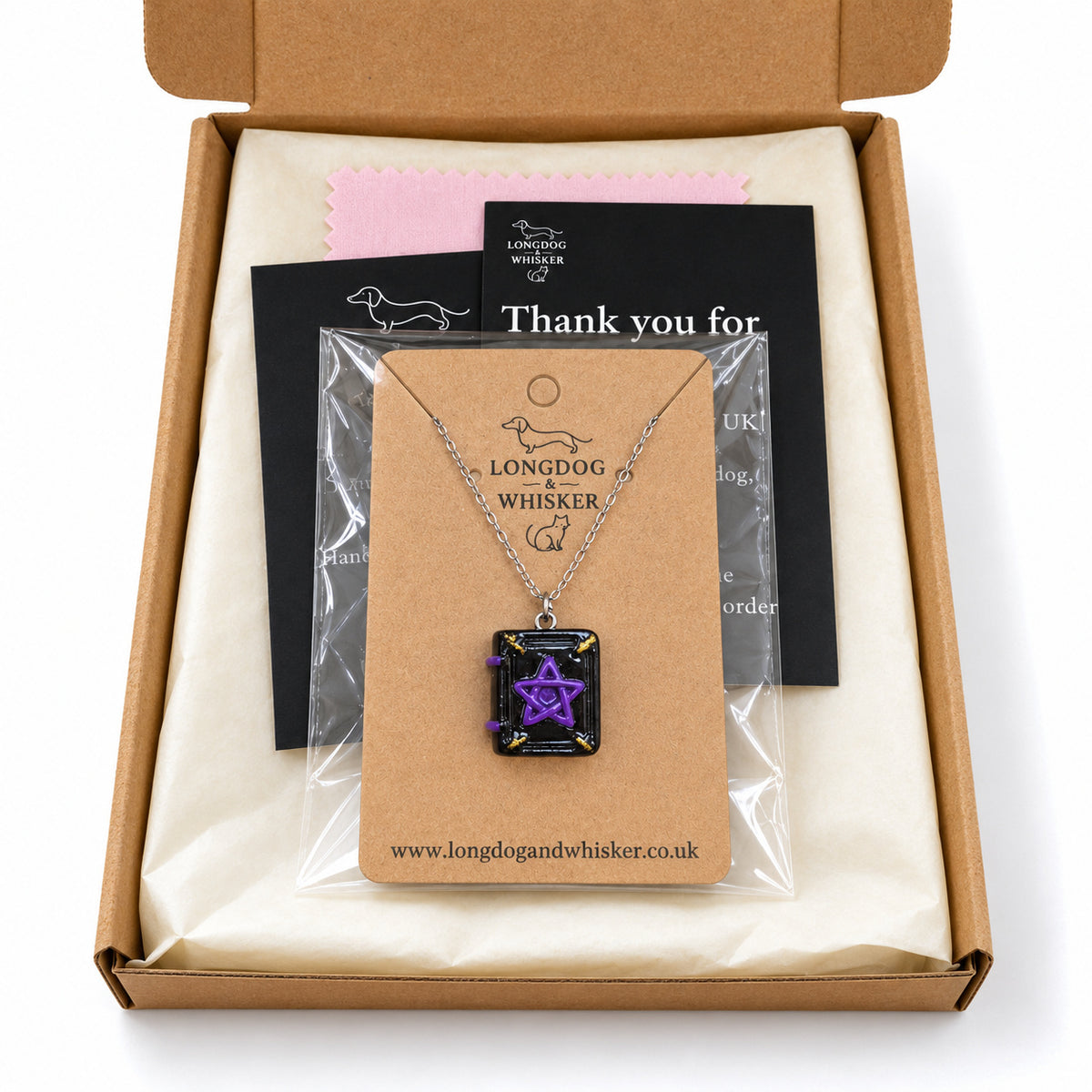 Spellbook necklace on its branded kraft card in a clear bag inside Longdog & Whisker kraft gift packaging