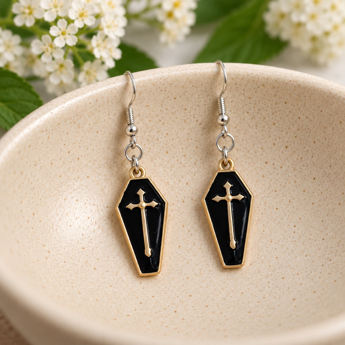 Black and gold-tone coffin cross earrings resting in a cream speckled ceramic bowl