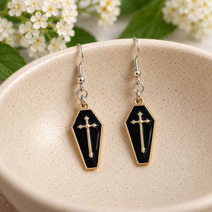 Black and gold-tone coffin cross earrings resting in a cream speckled ceramic bowl