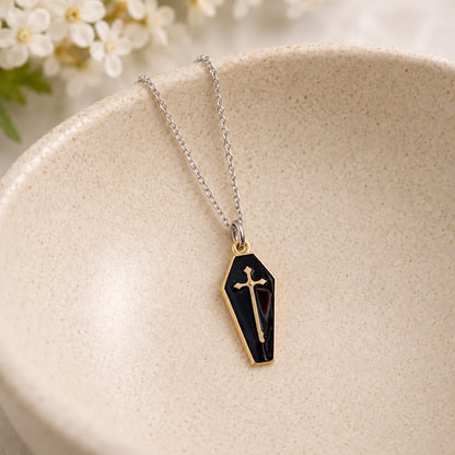 Black and gold-tone coffin cross necklace on a silver-tone chain resting in a speckled ceramic bowl