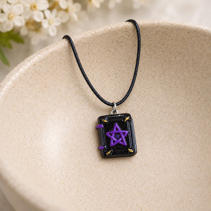 Black cord spellbook necklace with a purple pentagram resting in a speckled ceramic bowl