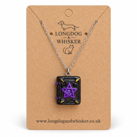 Black and purple spellbook necklace on a silver-tone chain on the Longdog & Whisker kraft card against white