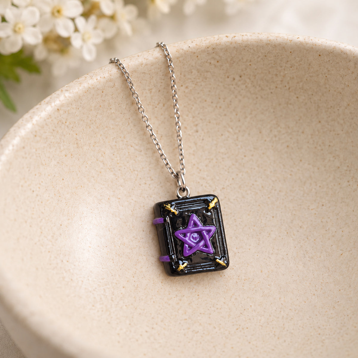 Black spellbook necklace with purple pentagram and silver-tone chain resting in a speckled ceramic bowl