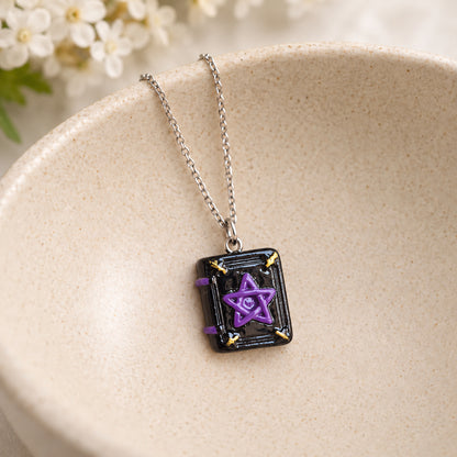 Black spellbook necklace with purple pentagram and silver-tone chain resting in a speckled ceramic bowl