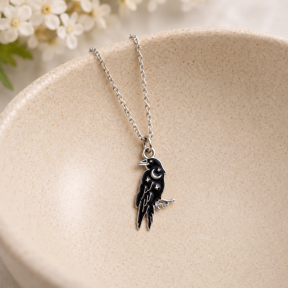 Black raven pendant and silver chain resting in a speckled ceramic bowl
