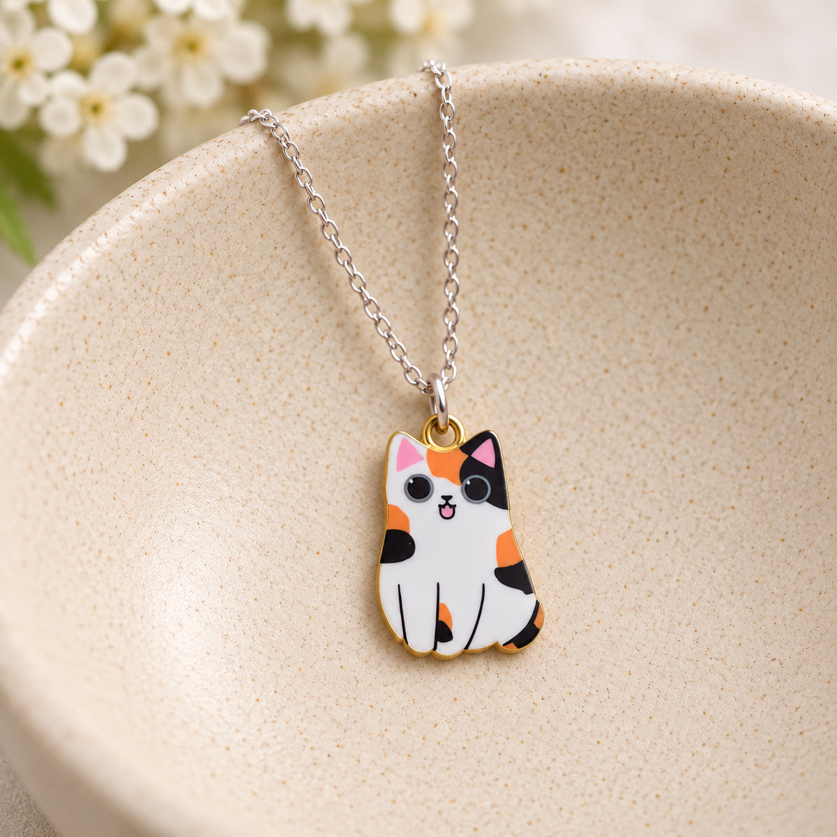 Orange, black and white calico cat pendant on a silver chain in a speckled ceramic bowl