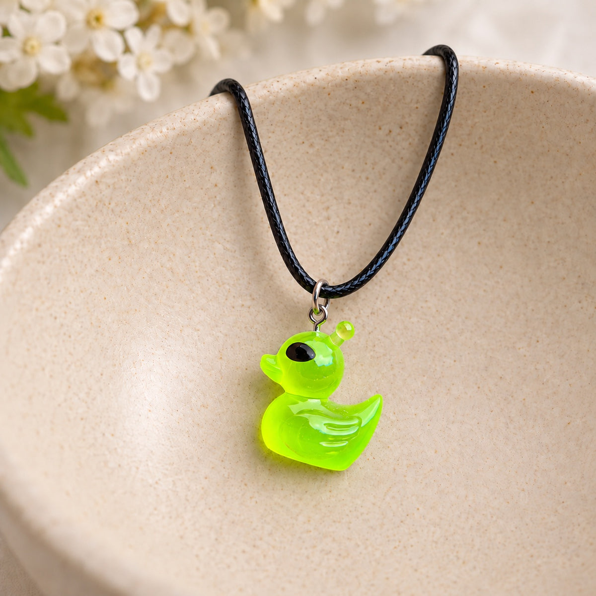 Translucent green alien duck necklace on a black cord resting in a ceramic jewellery bowl