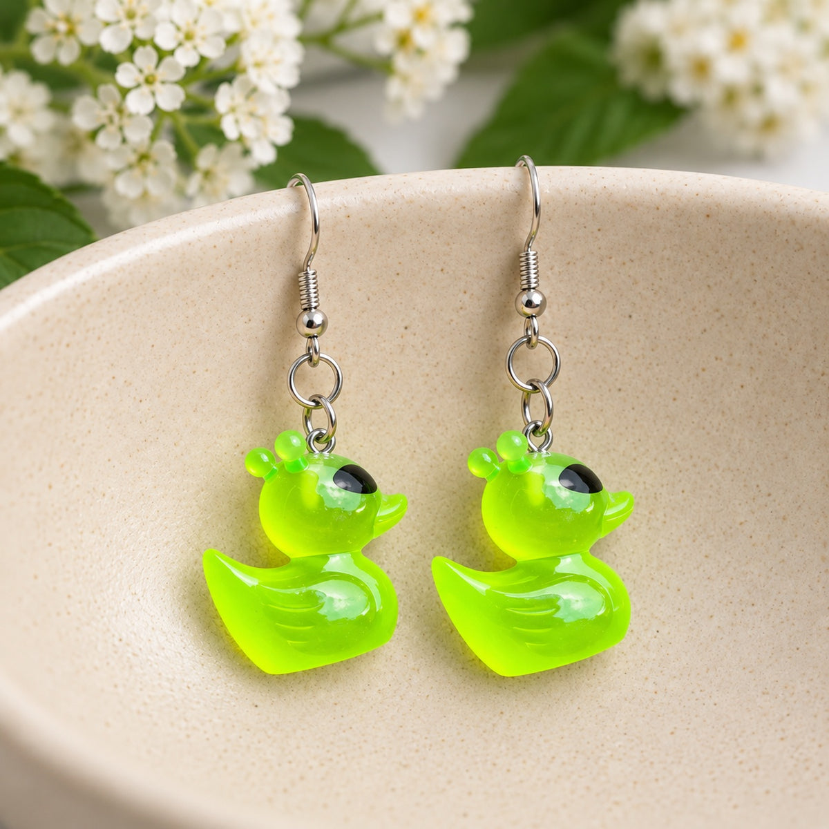 Green alien duck earrings with black eyes and rounded antennae resting in a speckled ceramic bowl