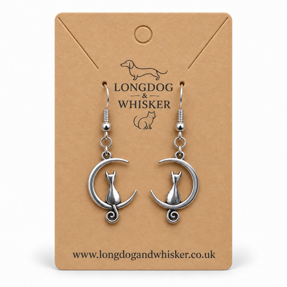Silver cat and crescent moon earrings, with the left tail in front of the moon and the right tail behind it, on a kraft display card.