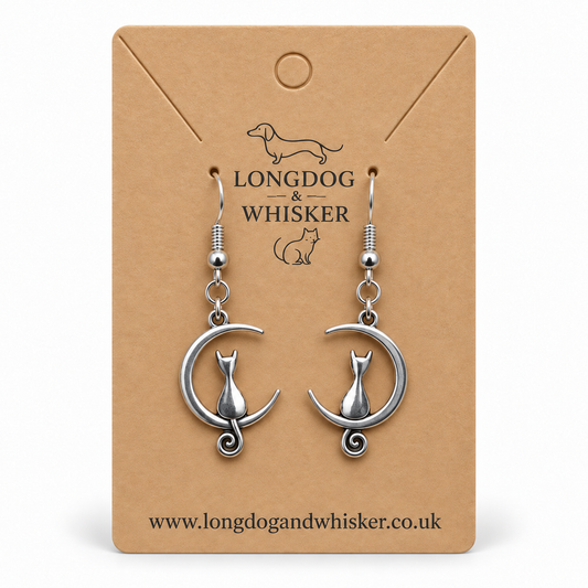 Silver cat and crescent moon earrings, with the left tail in front of the moon and the right tail behind it, on a kraft display card.