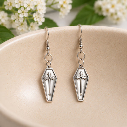 Silver coffin cross drop earrings resting in a speckled ceramic bowl.