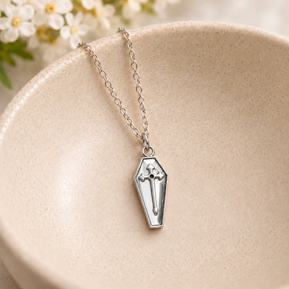 Silver coffin cross necklace resting in a speckled ceramic bowl.