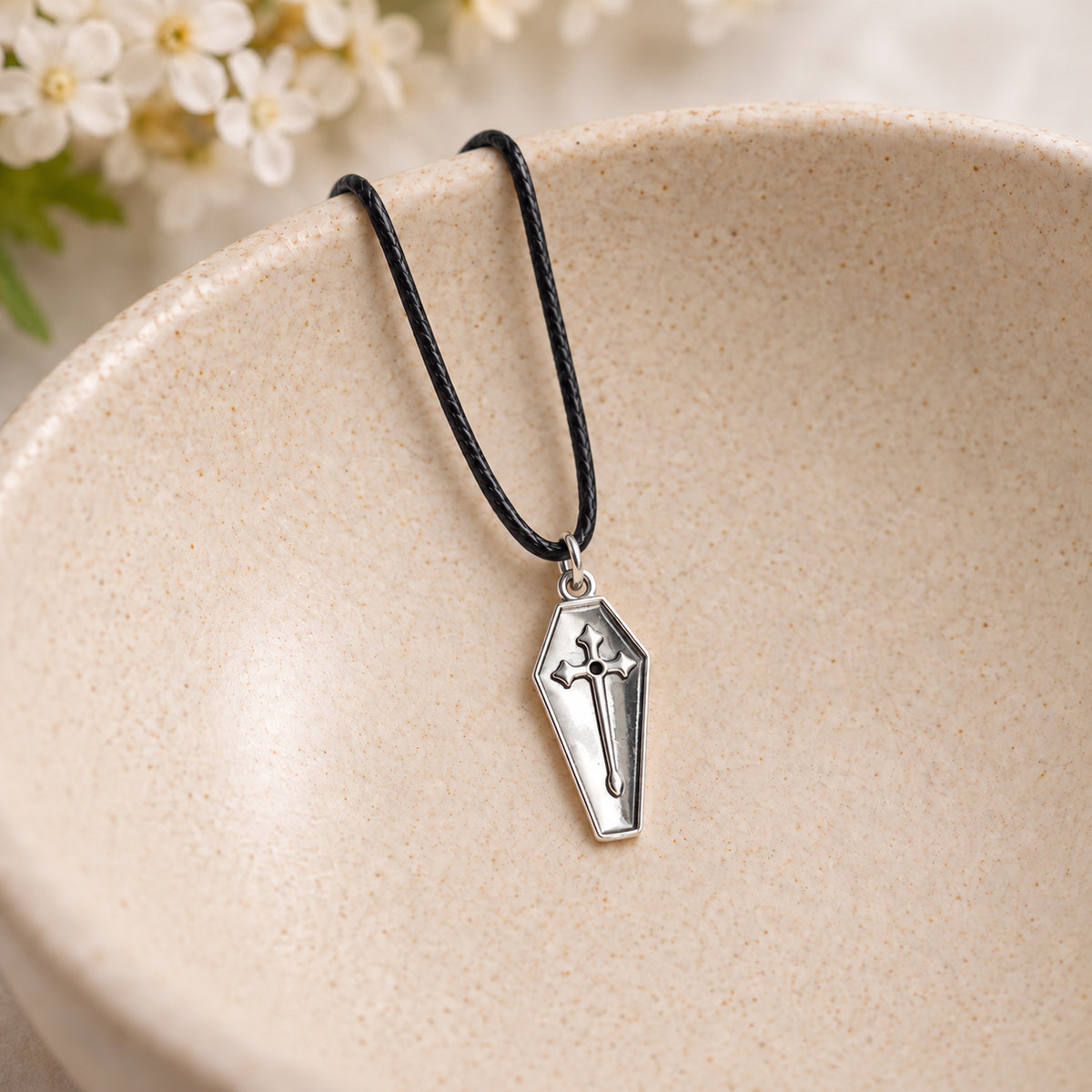 Silver coffin cross necklace on black cord resting in a speckled ceramic bowl.