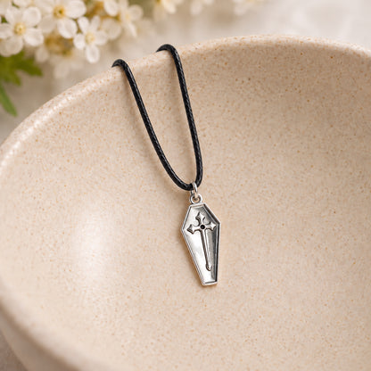 Silver coffin cross necklace on black cord resting in a speckled ceramic bowl.