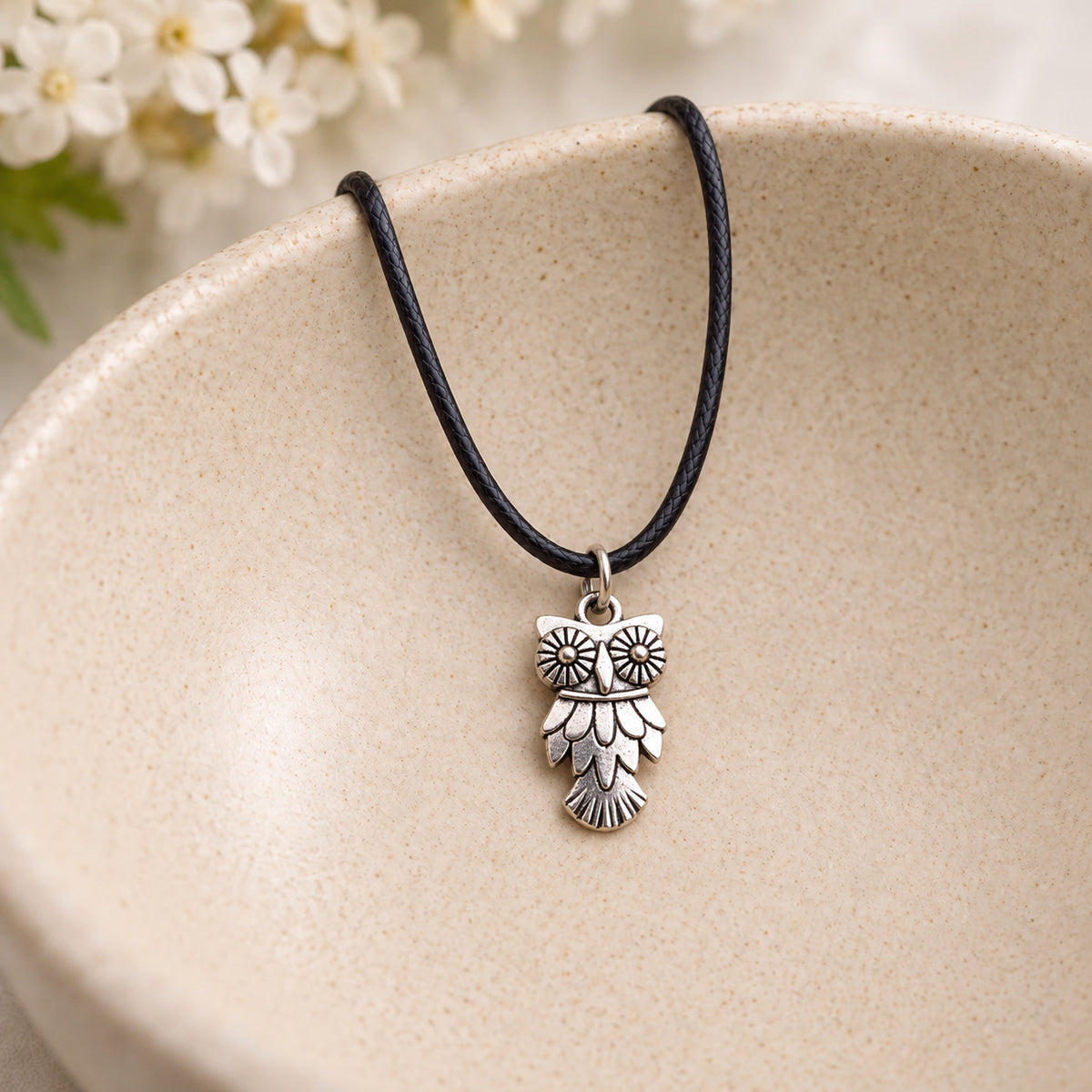 Silver-tone owl pendant on black cord resting in a speckled ceramic bowl