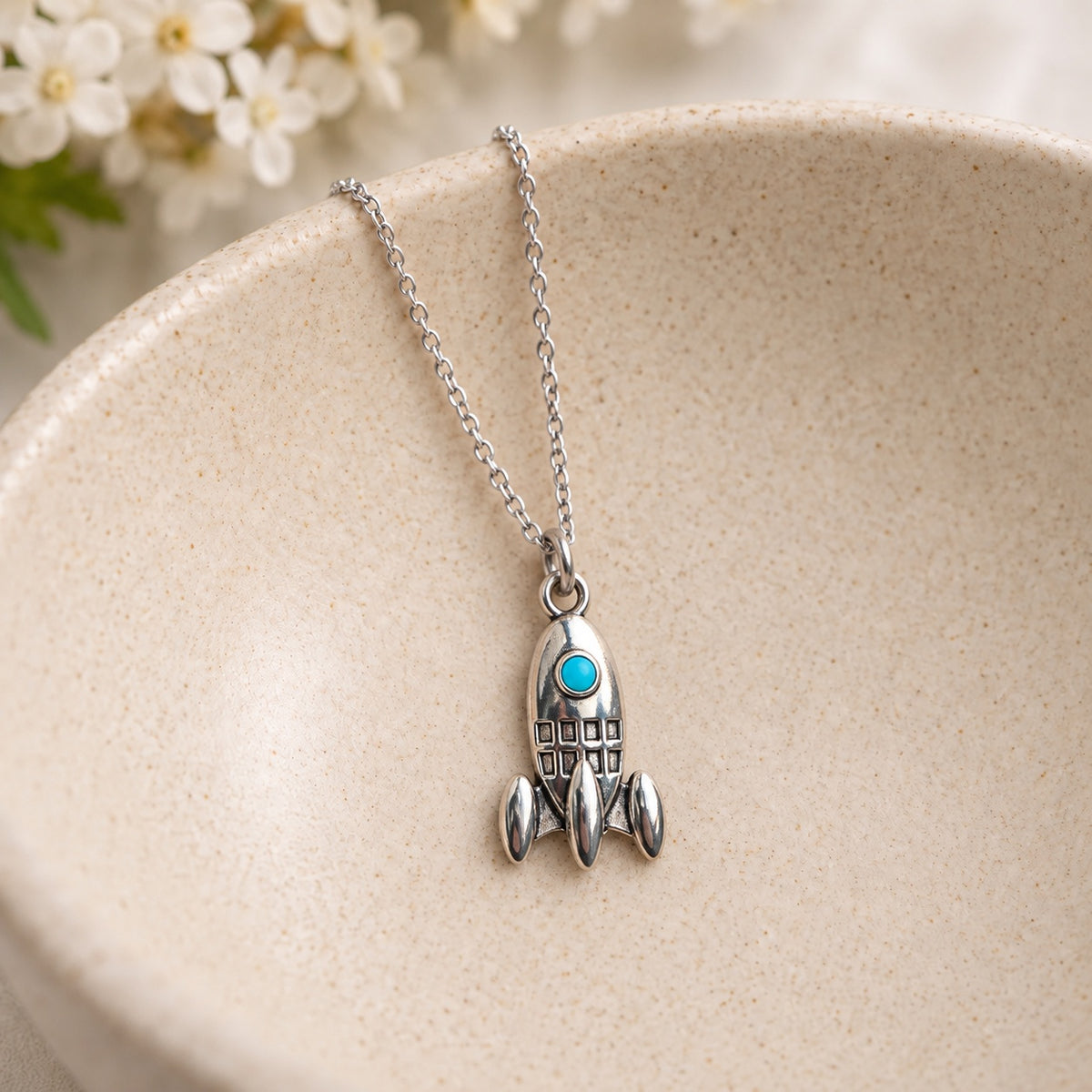 Silver-tone rocket necklace with turquoise porthole resting in a speckled ceramic jewellery bowl